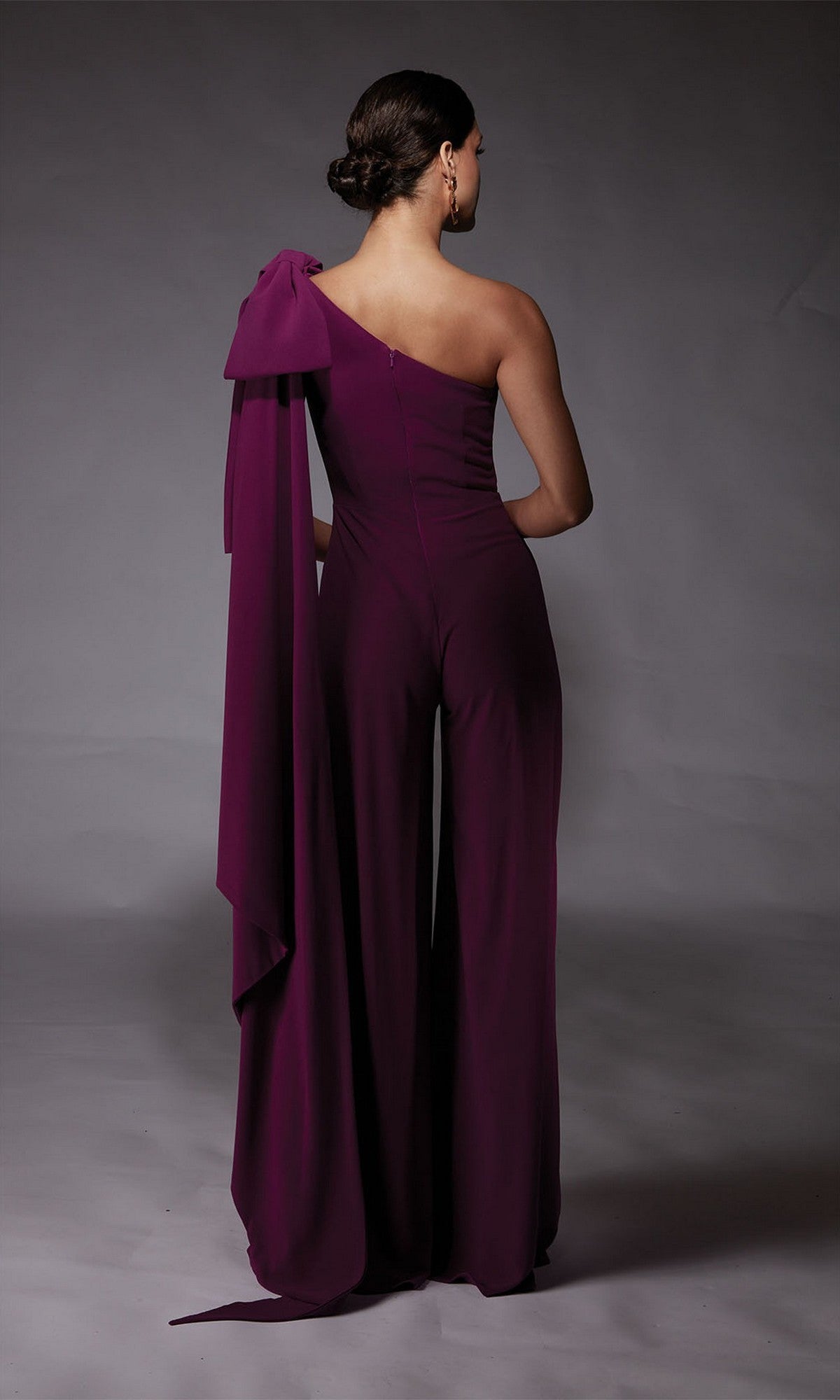 A woman faces away from the camera in the Alyce 70065 long formal jumpsuit—a deep purple, one-shoulder style with a dramatic draped bow, offering a bold alternative to a cocktail dress.